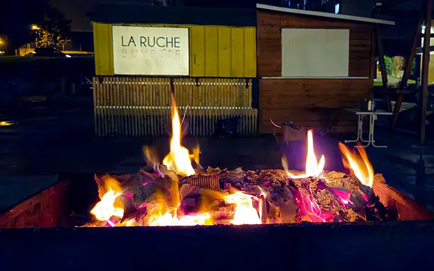 Un grand brasero brûle devant la buvette jaune La Ruche illuminée, un soir de mars 2025