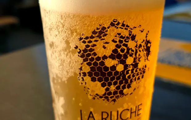 Gros plan sur un gobelet La Ruche Buvette, l’alvéole imprimée sur le verre, la place en arrière-plan flou, mai 2026